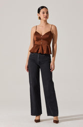 A woman is wearing a brown satin sleeveless top with thin spaghetti straps, button details down the front, and a peplum flare at the hem. She pairs it with high-waisted, straight-leg black jeans and brown pointed-toe heels. She also has gold earrings and rings on her fingers.